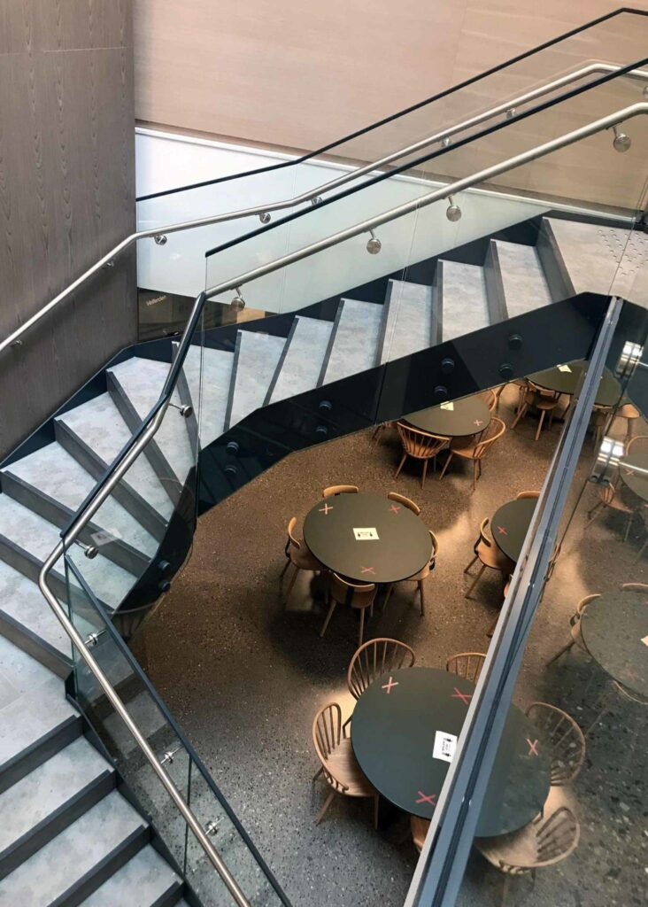 Moderne innendørs trapp med glassrekkverk og metallramme, sett ovenfra mot et sitteområde med runde bord.