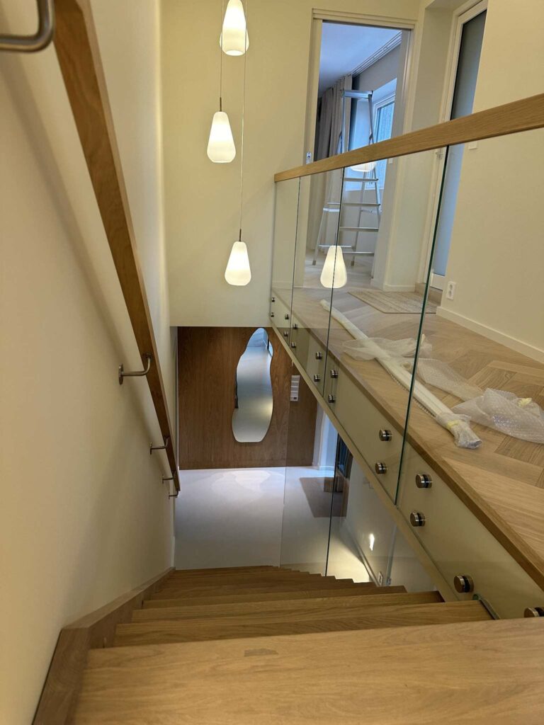 Eiketrapp sett ovenfra med glassrekkverk og hengende lamper.