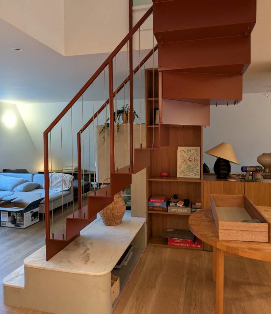 Sidevisning av en rødbrun ståltrapp montert over marmorpodium med bokhylle og stue i bakgrunnen.