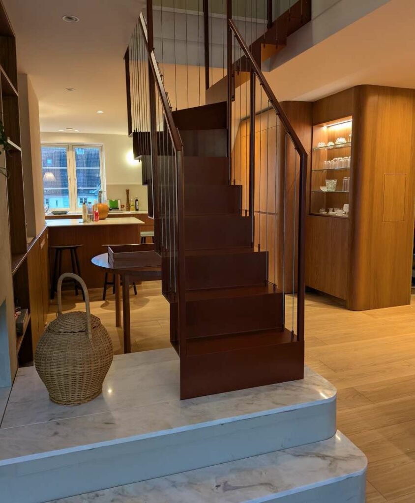 Rødbrun ståltrapp som står på et marmorpodium i et moderne kjøkken- og oppholdsrom.