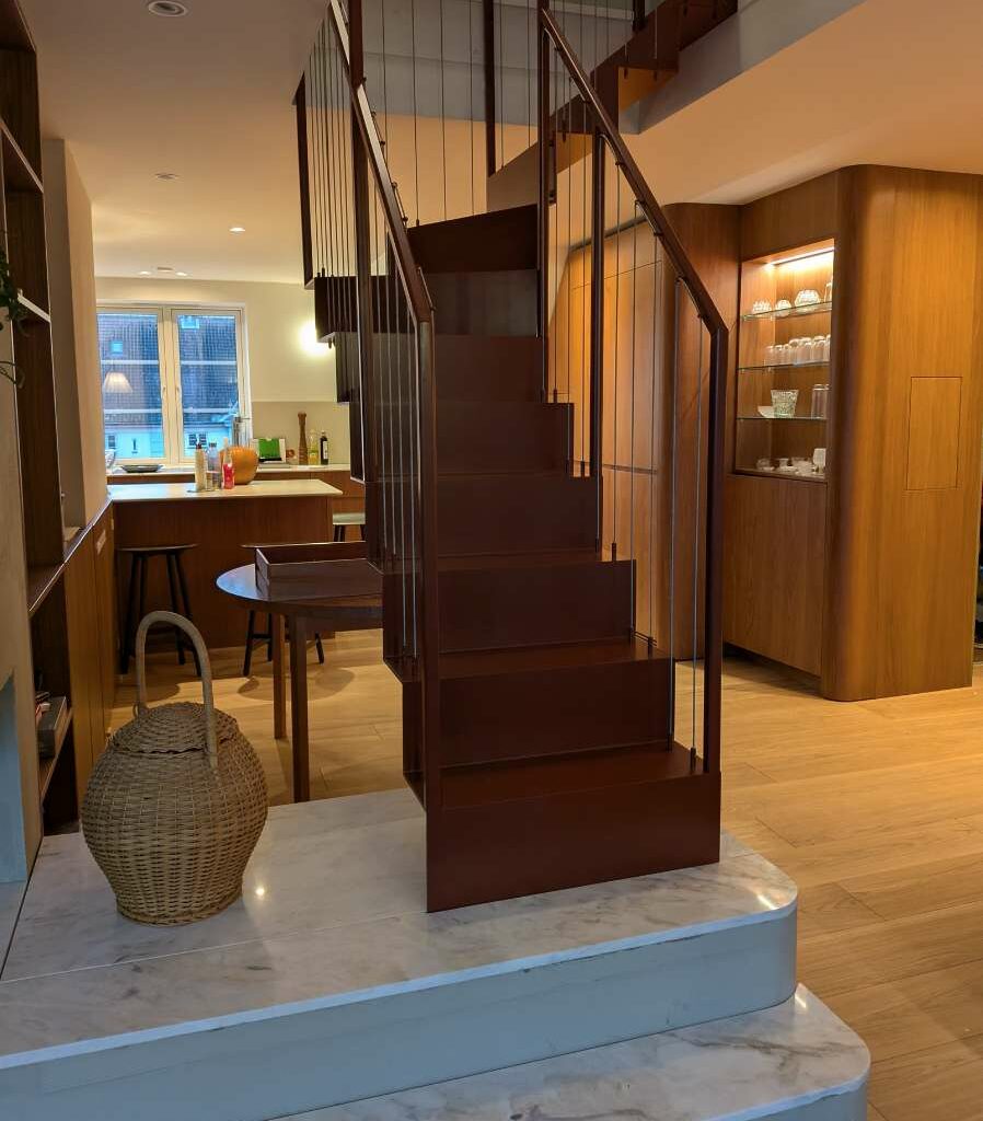 Rødbrun ståltrapp som står på et marmorpodium i et moderne kjøkken- og oppholdsrom.