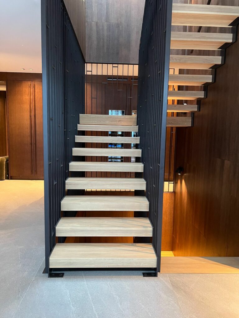 Trappeløp med eiketrinn og sort rekkverk sett ovenfra, omgitt av mørke trevegger og varm belysning.