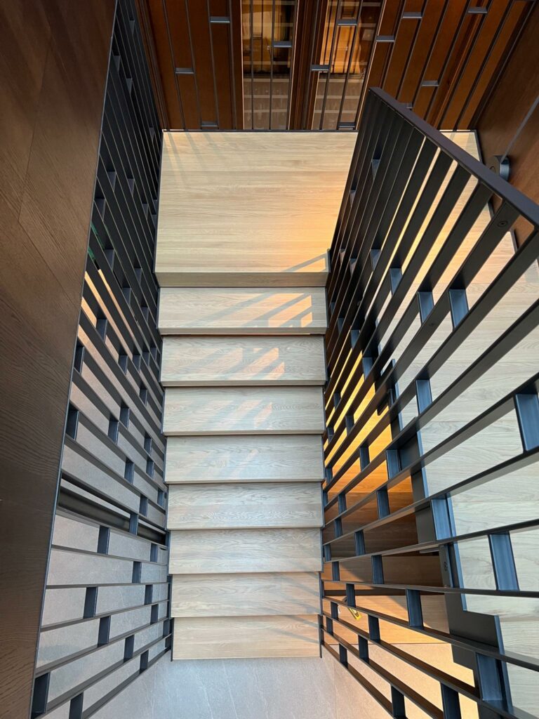 Oversiktsbilde av moderne innvendig trapp med lyse eiketrinn og dekorativt metallrekkverk.